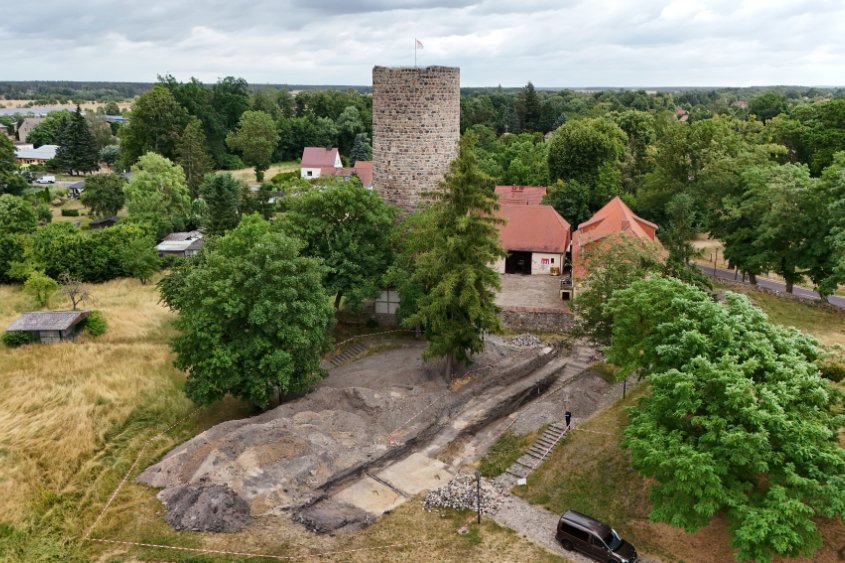 Schrägluftbild des Grabungsschnitts im westlichen Bereich der Burg Loburg; Blickrichtung Osten. Im Westen des Grabungsschnitts ist der helle anstehende Boden zu erkennen, daran anschließend sind die dunklere Verfüllung des frühmittelalterlichen Befestigungsgrabens und Wallschichten zu sehen; oberhalb der Grabungsfläche befindet die jüngere Burganlage mit dem Bergfried. © Landesamt für Denkmalpflege und Archäologie Sachsen-Anhalt, Anja Tuschwitz.